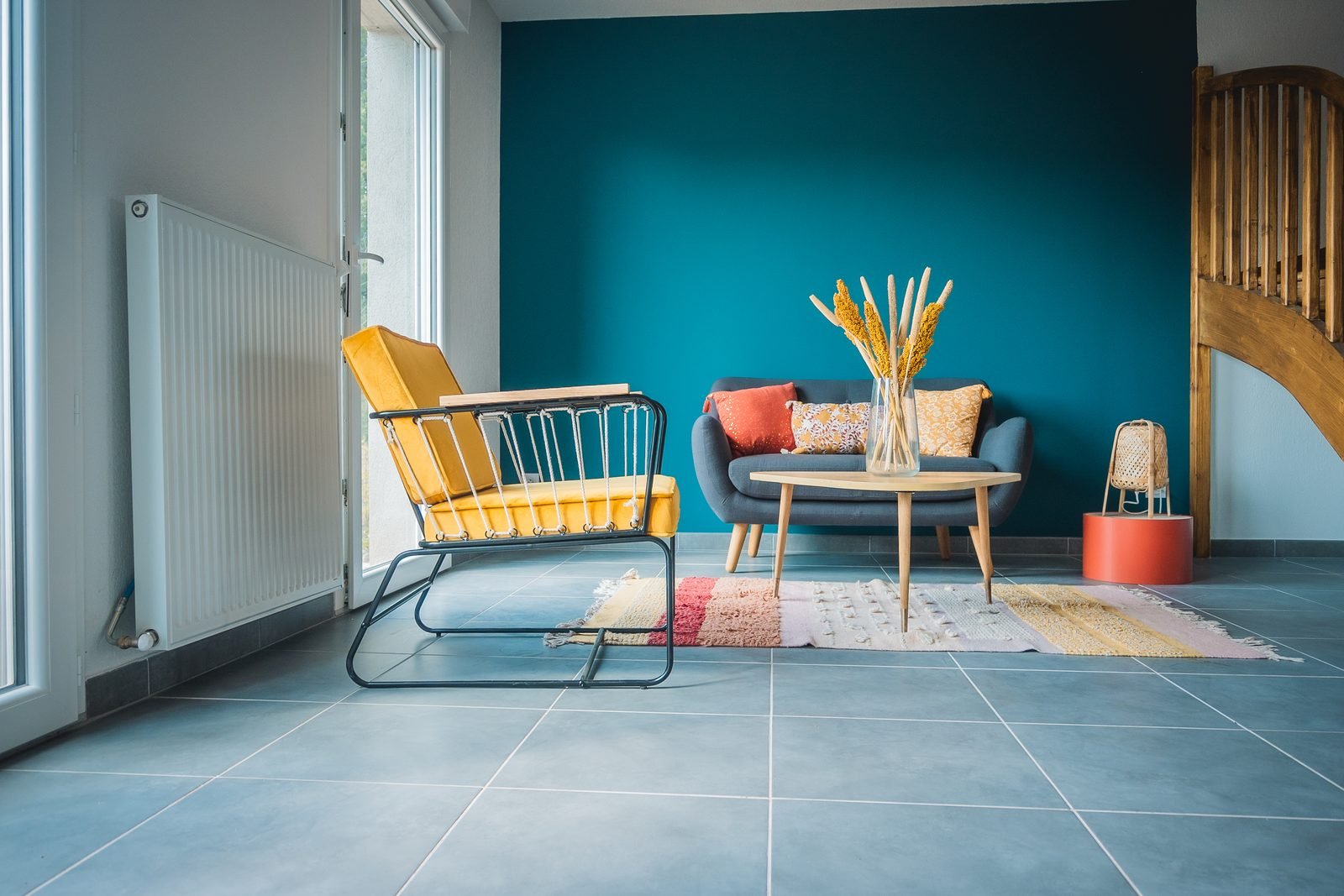 Sala com parede azul-petróleo, sofá azul, poltrona mostarda e tapete colorido