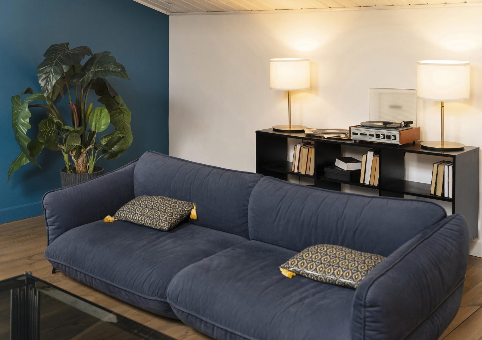 Sala com parede azul-petróleo, sofá azul-marinho, estante preta com livros e abajures acesos
