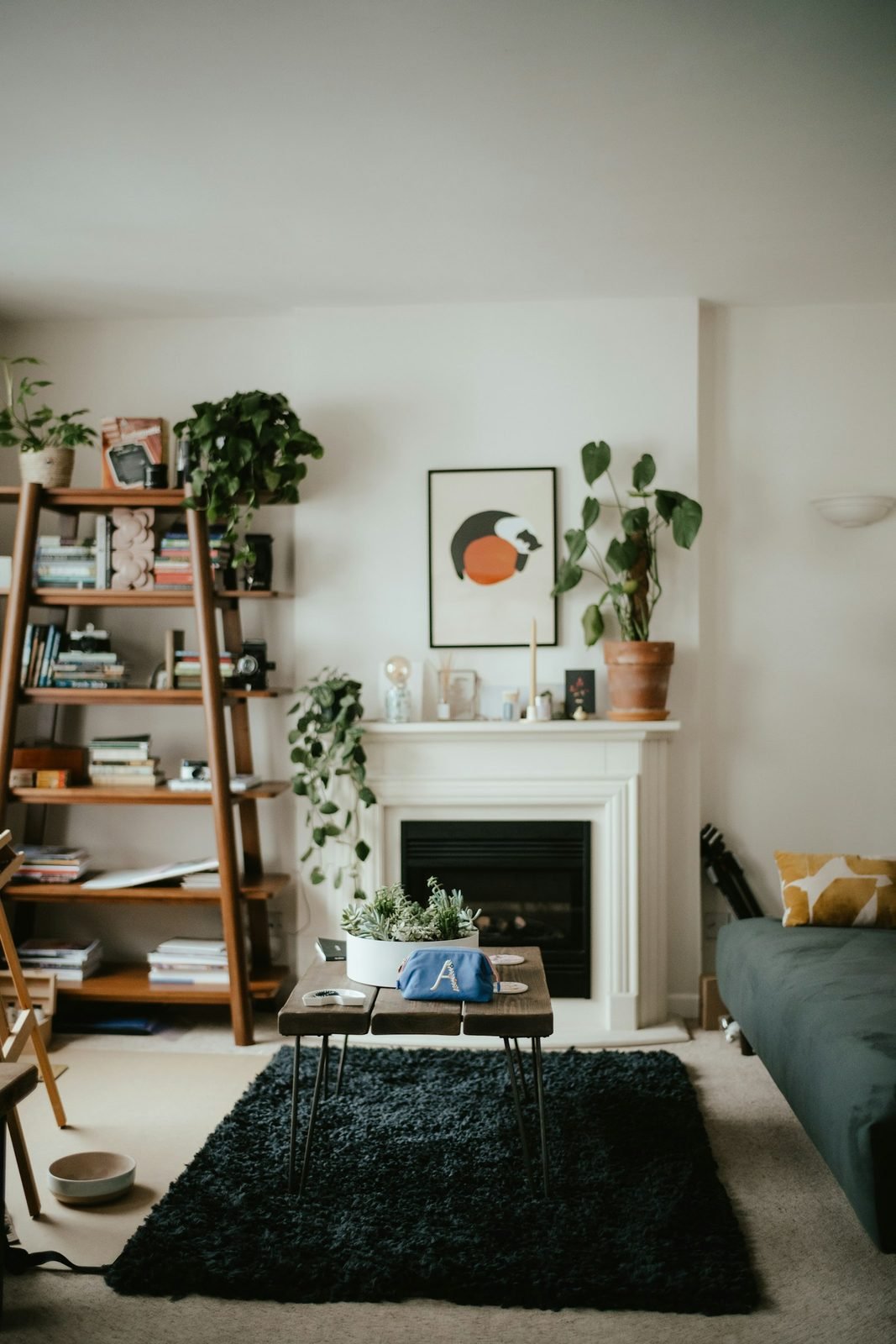 Sala com estante de madeira em formato de escada cheia de livros e plantas, lareira branca, quadro e tapete preto sob mesa de centro