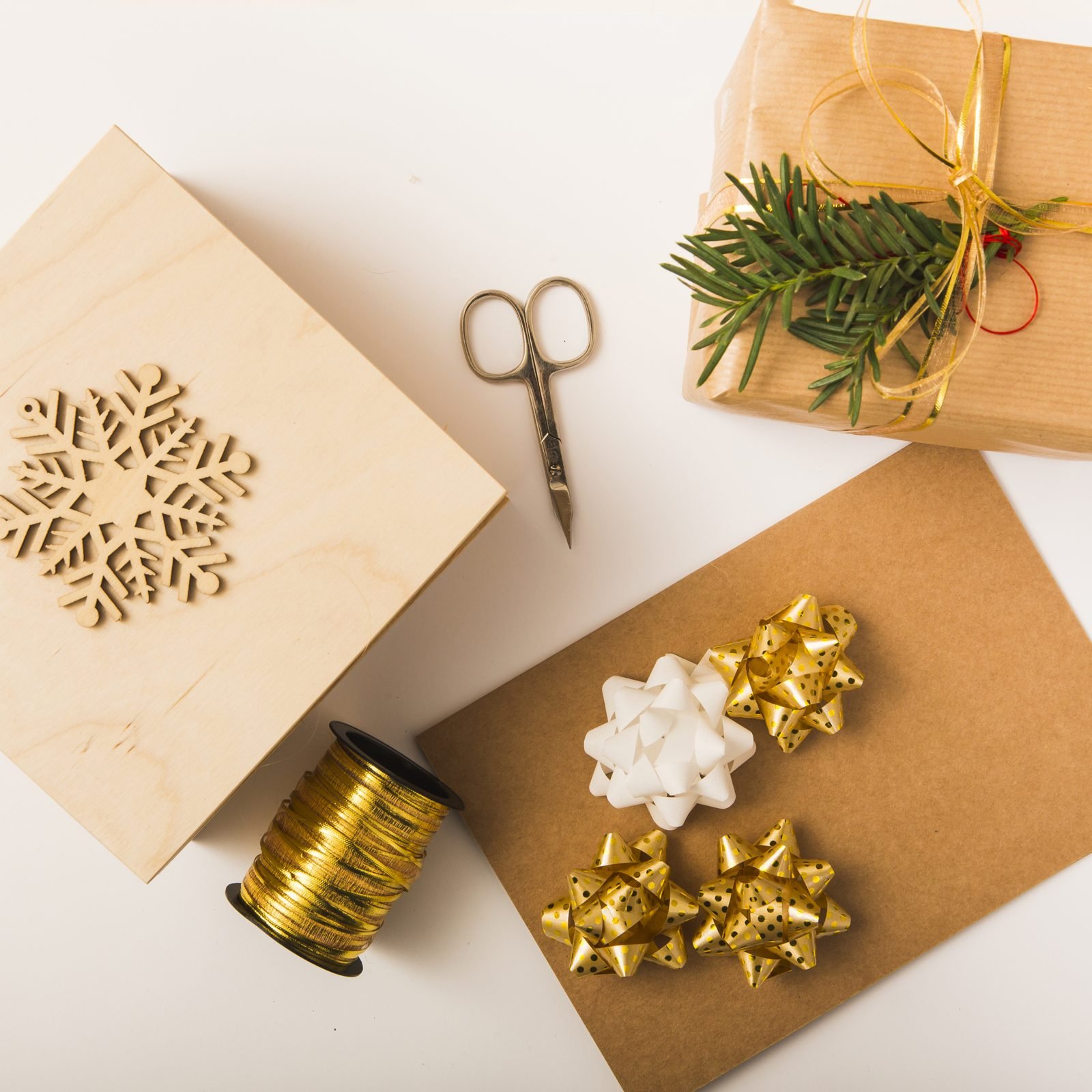 Kraft paper, scissors, gold bows, ribbon and a wooden snowflake on a white table