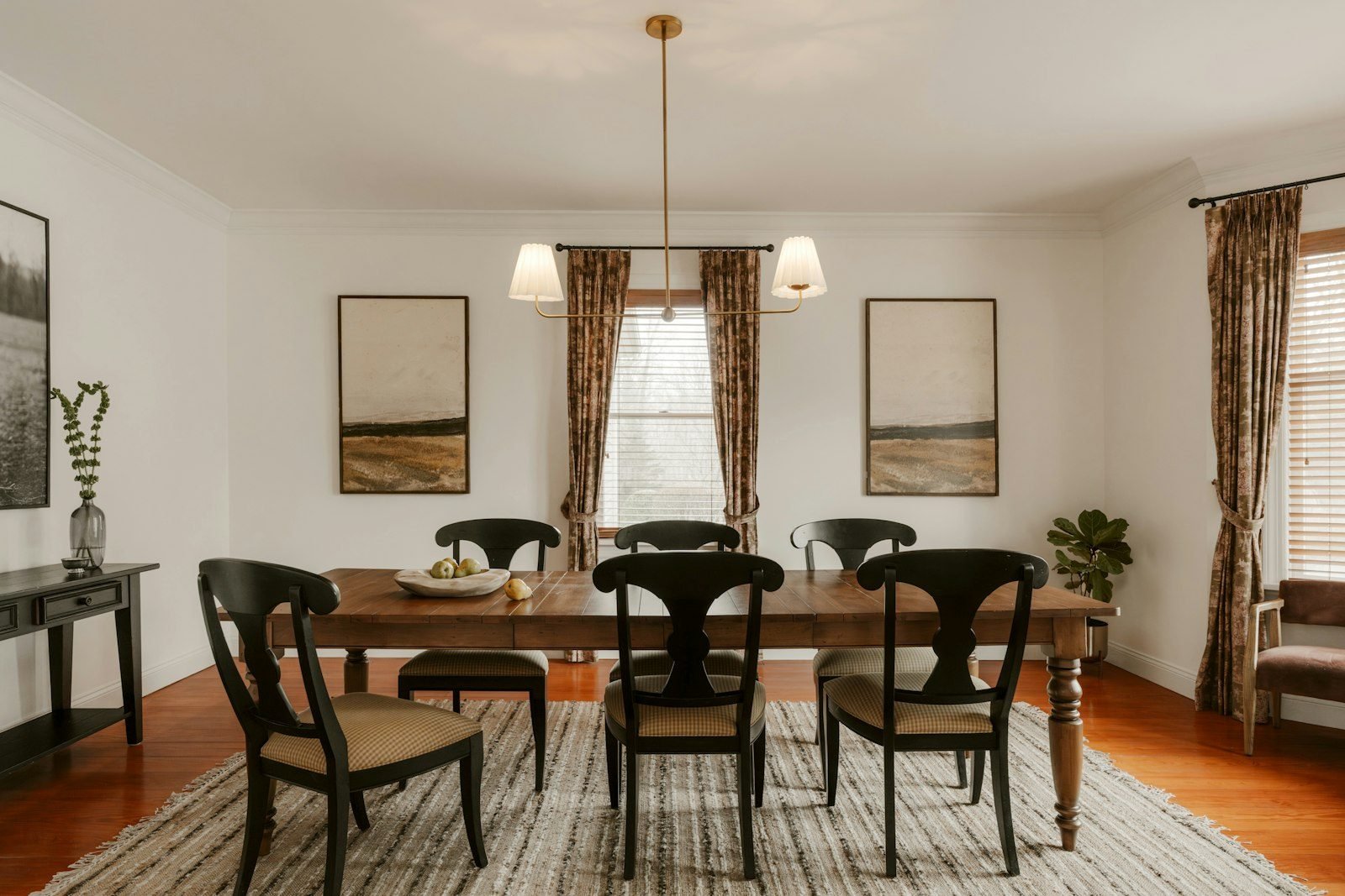 Sala de jantar com mesa de madeira, cadeiras pretas, tapete listrado, pendente dourado e aparador escuro na lateral