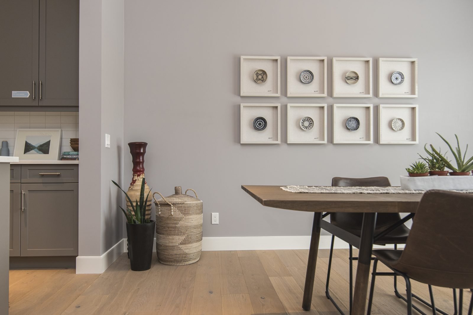 Parede cinza com composição de oito quadros brancos com pratos decorativos ao lado de mesa de jantar de madeira e cesto de fibra