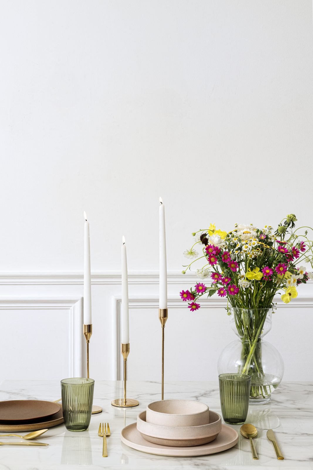 Mesa de jantar com tampo claro, castiçais dourados com velas brancas, vaso de vidro com flores do campo e louças em tons rosados