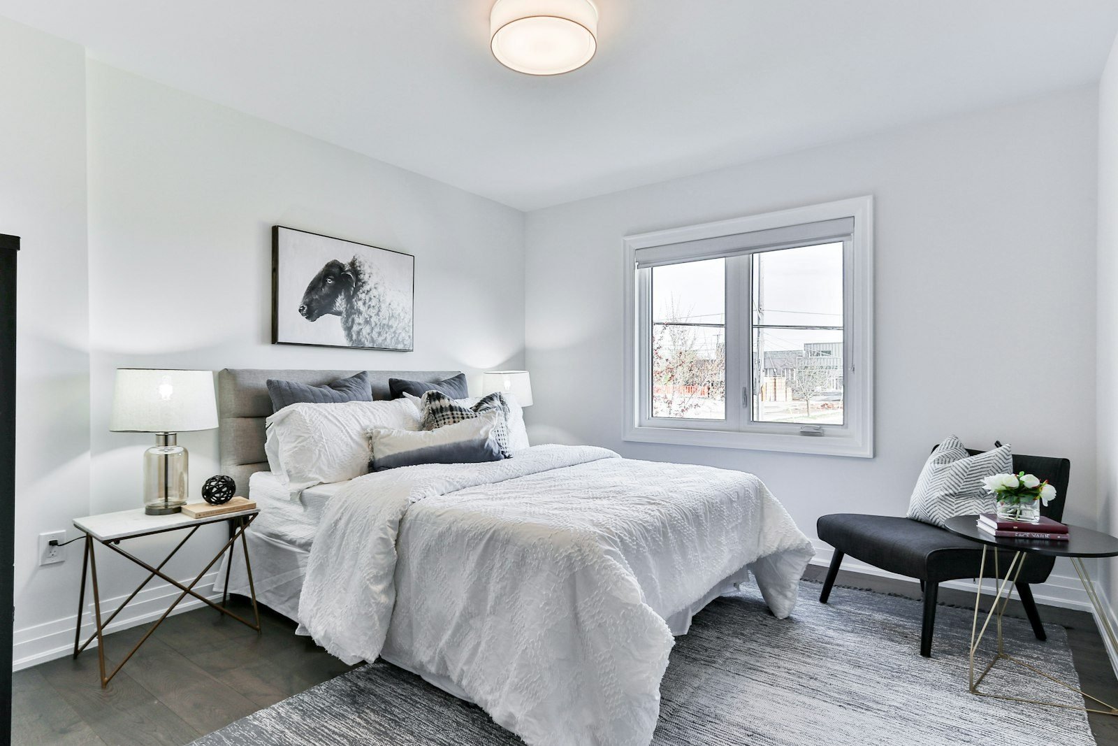 Quarto claro com cama de casal com roupa de cama branca, almofadas cinza, abajures e poltrona escura perto da janela