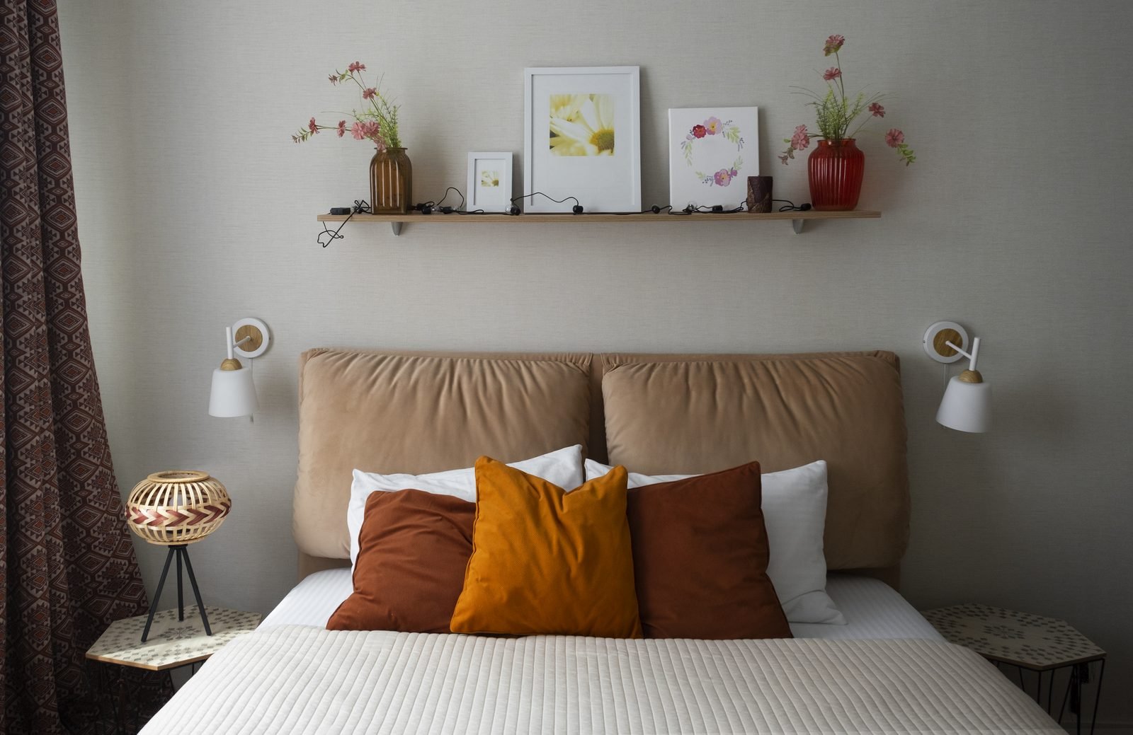 Cama de casal com cabeceira estofada caramelo, almofadas laranja e terracota, arandelas brancas e prateleira com quadros