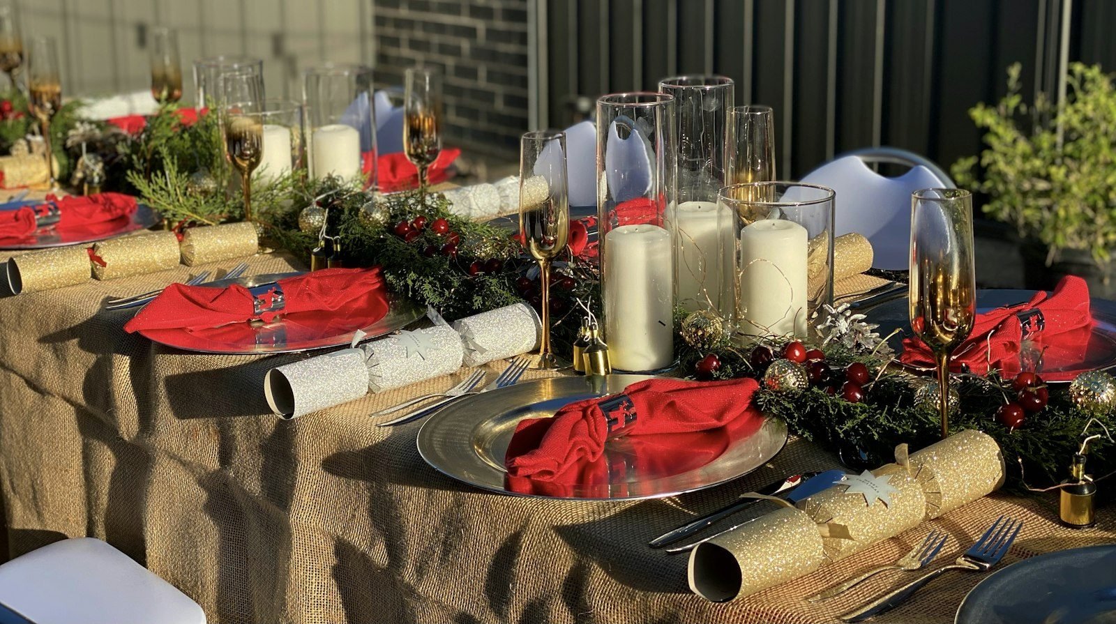 Christmas table with a burlap tablecloth, red napkins, white candles in glass cylinders and green branches down the center