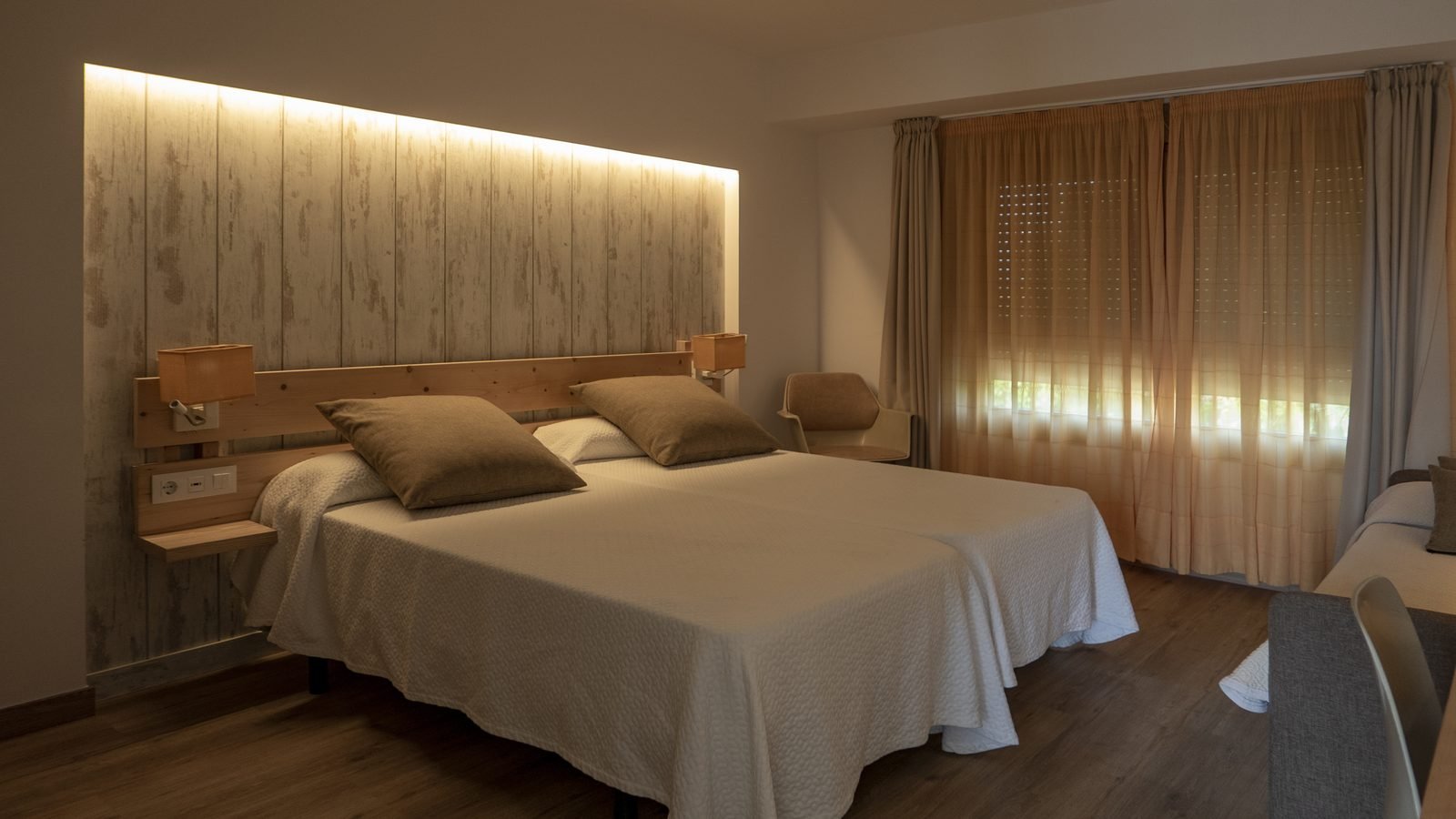Quarto com fita de LED embutida acima da cabeceira de madeira, luminárias de leitura e cortinas claras